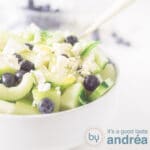 A square photo showing part of a white bowl filled with blueberry melon salad with feta and cucumber on a white background.