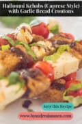 A portion of a plate of kebabs with halloumi cherry tomatoes, basil, garlic bread, and balsamic vinegar. A caption reads: Halloumi Kebabs (Caprese Style) with Garlic Bread Croutons