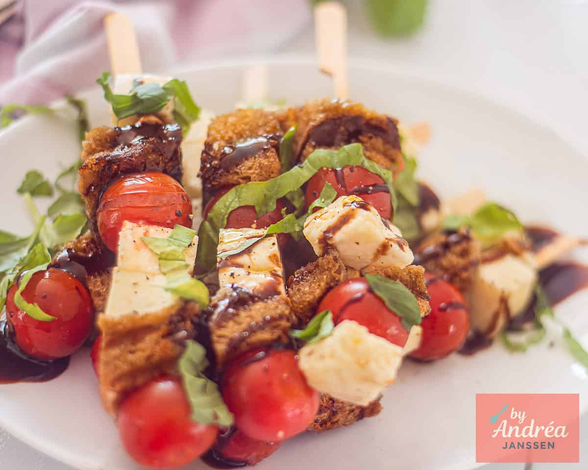 A white dish with Halloumi skewers drizzled with balsamic cream with tomatoes, basil and garlic bread