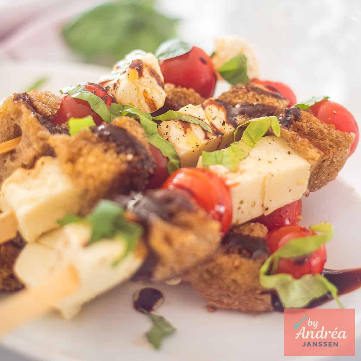 A square photo of a plate of skewers with halloumi, cherry tomatoes, garlic bread, basil and balsamic cream