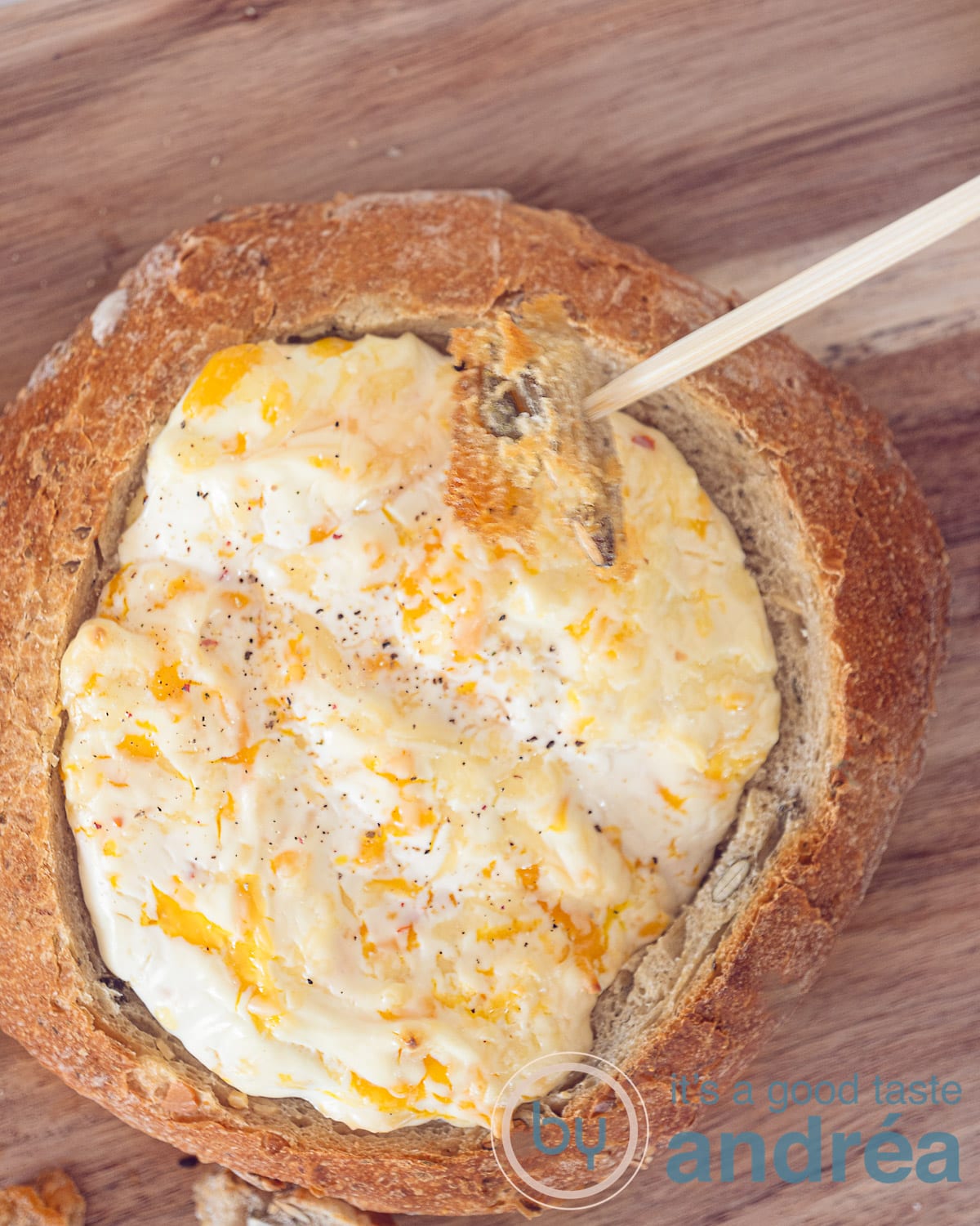 A photo from the top with a bread bowl filled with four cheese fondue. A piece of bread is dipped in the sauce. A wooden plate in the back.