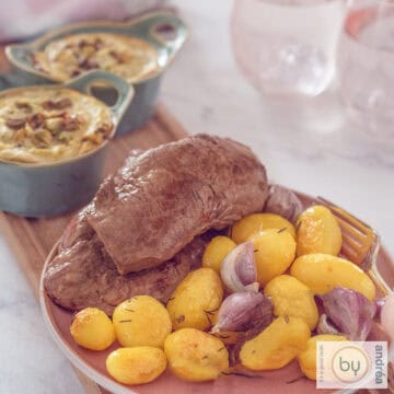 A square photo with a pink plate with potatoes, roasted garlic and sirloin steak. However, there are two bowls with broccoli goat cheese quiche