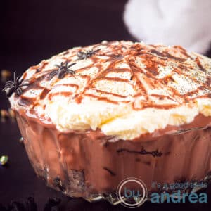 A square photo showing part of a glass bowl filled with oreos, brownies, chocolate custard and whipped cream. On top of a caramel spider web with spiders.