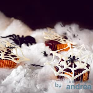 a square photo of nutella cupcakes with white chocolate spiderwebs on a white cloudy ground.