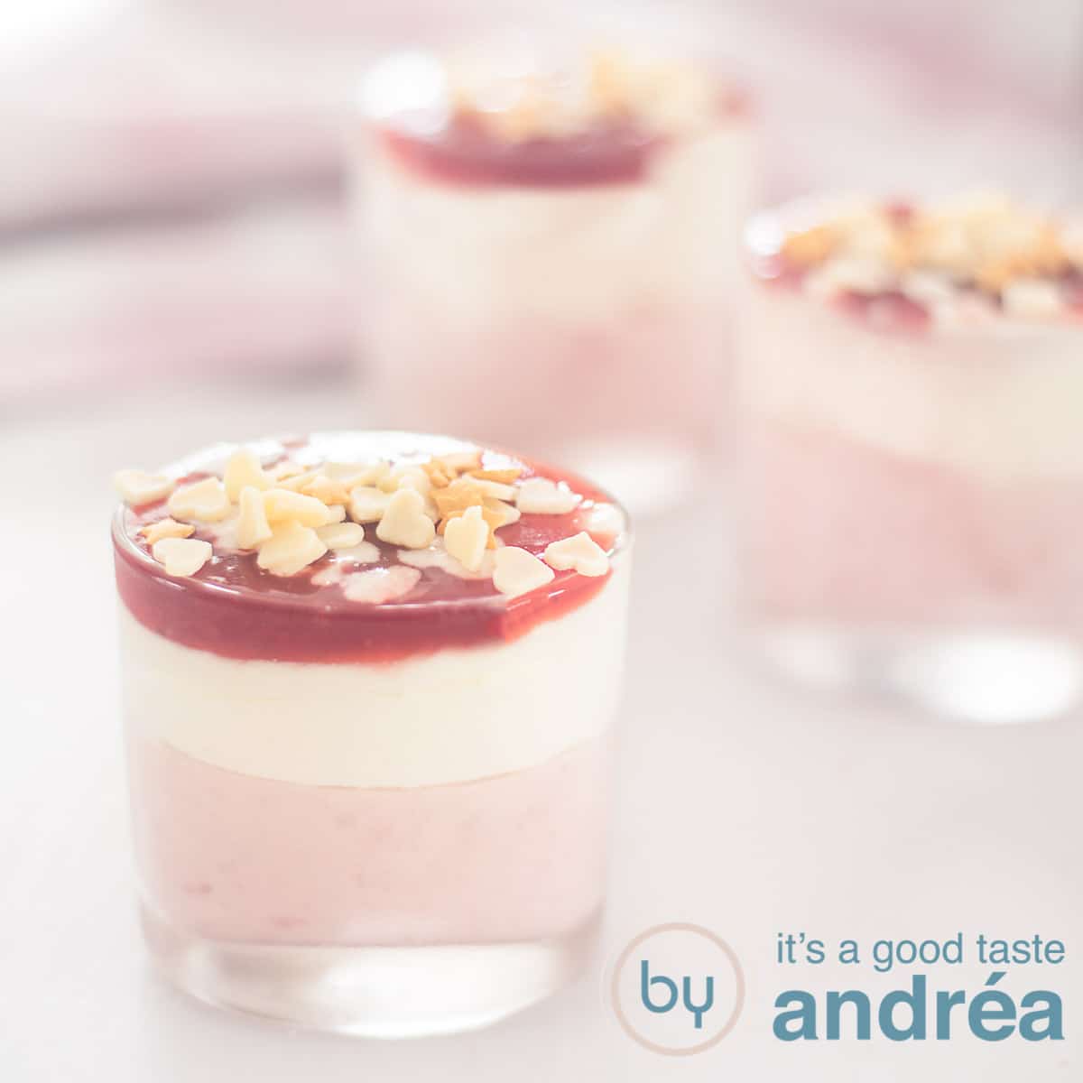 A square photo with three dessert glasses filled with strawberries with white chocolate mousse and a glaze. A pink white towel in the background