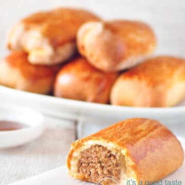 Half a Dutch worstenbroodje (sausage rolls) on a white napkin on a white backdrop. More Worstenbroodjes in the back at a white plate.