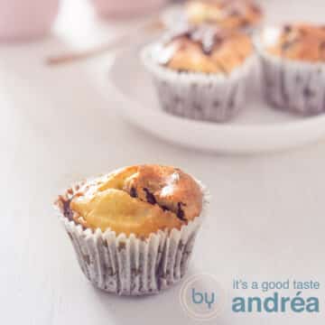 A square photo with a chocolate cheesecake cupcakes in the front left and a plate with three cupcakes in the back right. Everything on a white background.