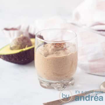 A square photo with a glass of chocolate coconut smoothie. An avocado and a pink white tea towel in the background.