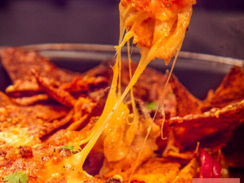 A square picture of a casserole full of nacho's. One is pulled up and got cheesy strings in between.