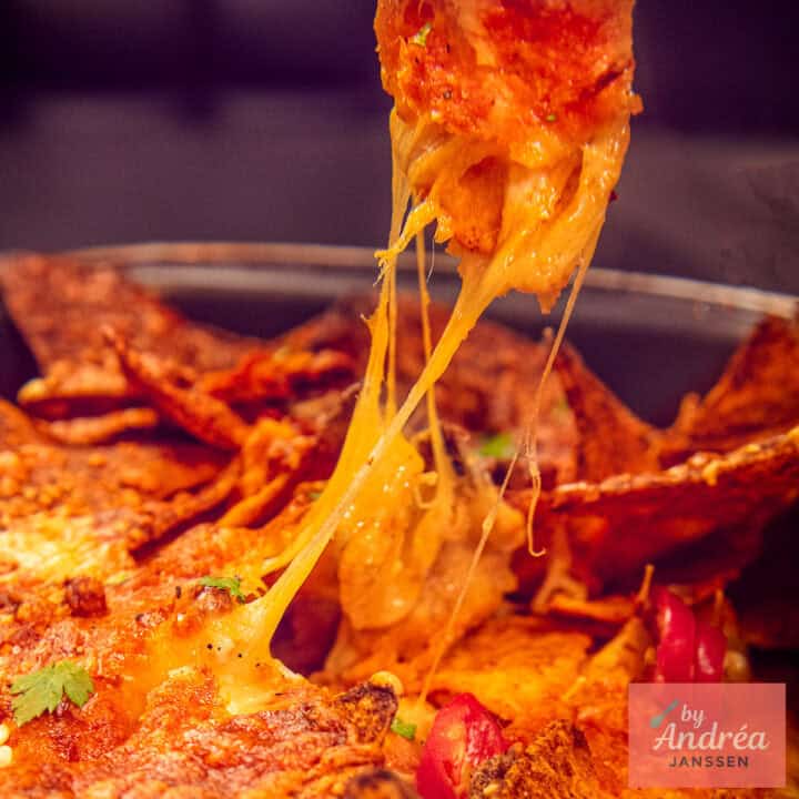 A square picture of a casserole full of nacho's. One is pulled up and got cheesy strings in between.