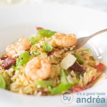 A square photo with Orzo, shrimp, tomato and zucchini on a white plate. A golden fork scoops up a shrimp.