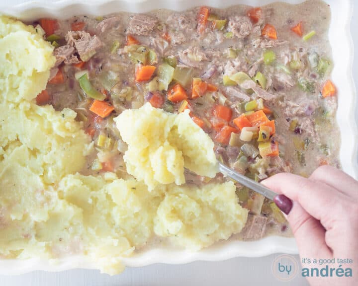 Mashed potatoes are spooned over the vegetable filling with tuna in a white baking dish.
