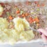 Mashed potatoes are spooned over the vegetable filling with tuna in a white baking dish.