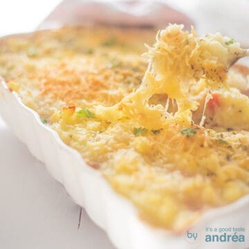 A square photo with an oven dish filled with potato casserole with tuna. A spoon takes a bite of the cheesy top.