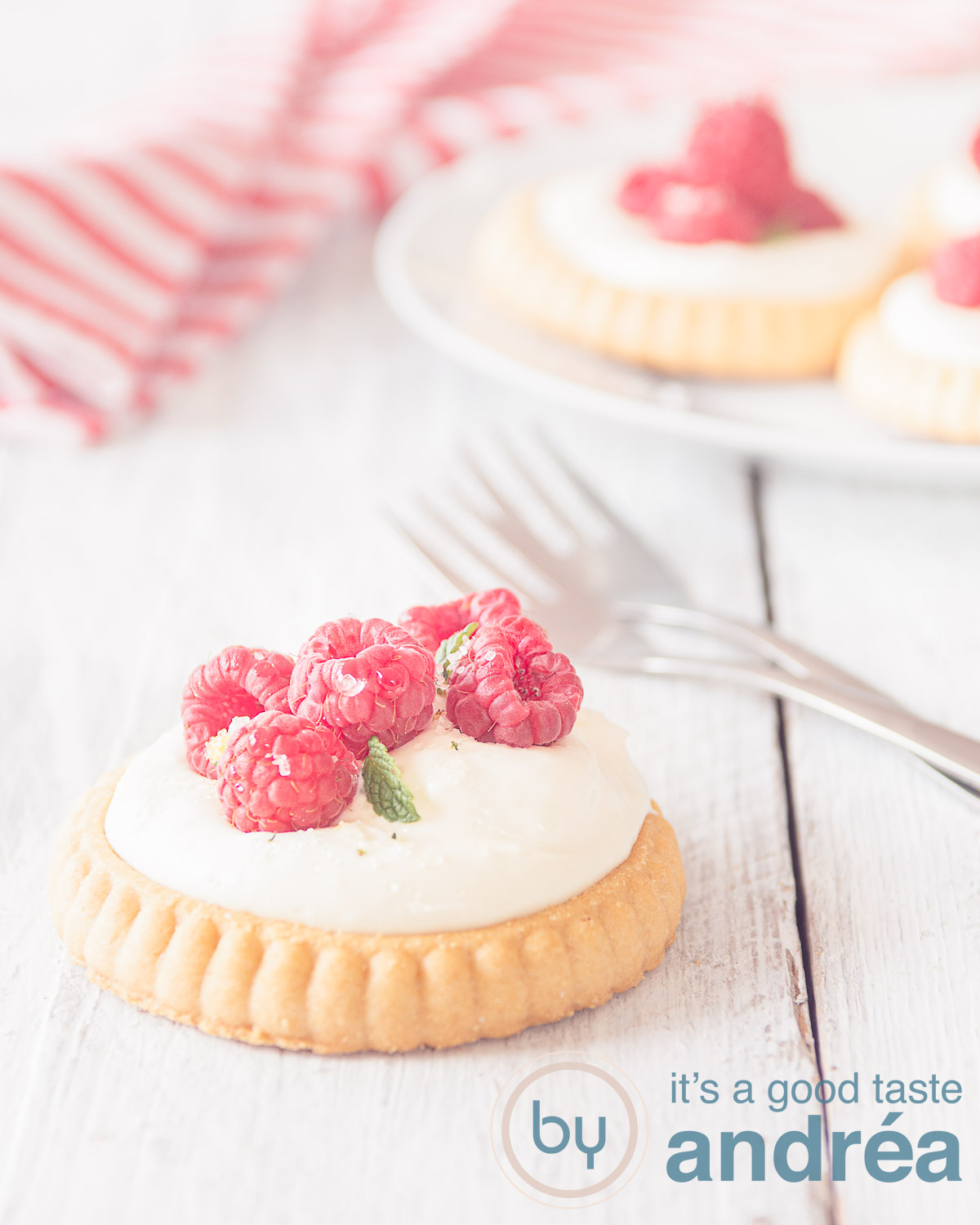 Raspberry tartlet with mascarpone cream on a white wooden background. Two forks behind the first tartlet. A red white striped cloth in the back.