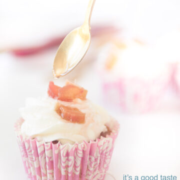 A pink cupcake wrapper filled with a cupcake with rhubarb, cream topping and rhubarb pieces. A white background