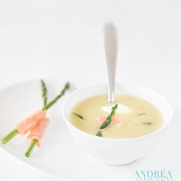 a white plate with a bowl of mustard soup and two green asparagus and a bit of smoked salmon