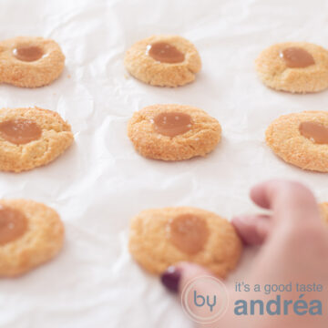 a square picture with eight coconut caramel cookies on a white baking paper. A hand is grabbing the middle one.
