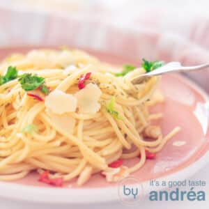 A square photo showing part of a pink plate, a mountain of spaghetti seasoned with pepper, olive oil and garlic, parsley and sheared parmesan cheese