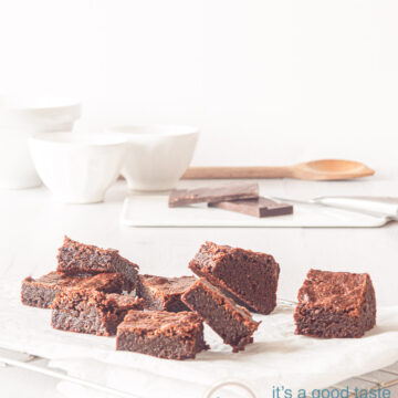 pieces of brownies scattered on a piece of baking paper. In the background some bowls, chocolate and wooden spoon
