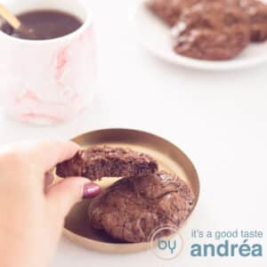 A square photo with a Brownie cookie, a hand takes it off the tray. In the back left is a pink mug with coffee. At the top right a white board with a number of brookies. A white background