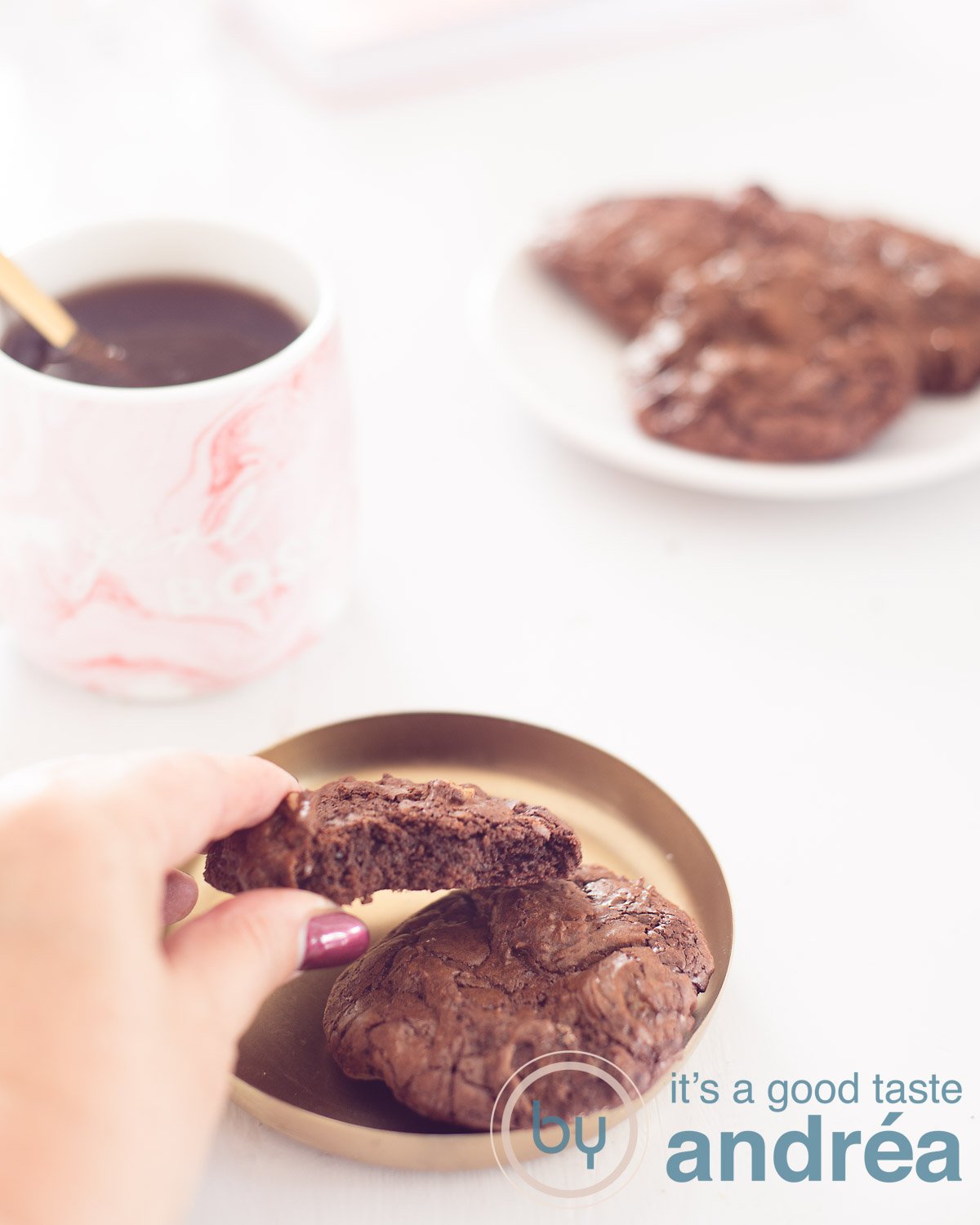 Brownie Walnut Biscuits on a golden platter. A hand takes it off. In the left background a pink mug with coffee. A sign with more brookies in the back right. A white background.