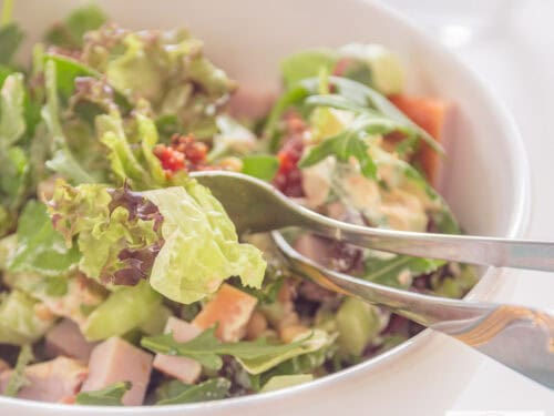 A square photo of a white bowl with green salad, smoked chicken and sun-dried tomatoes.