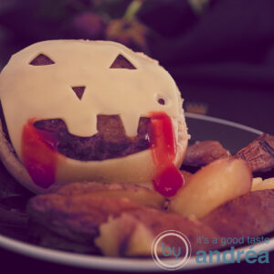 a square photo of a cheeseburger with a Jack-O-Lantern face and oven-baked fries on a black plate.