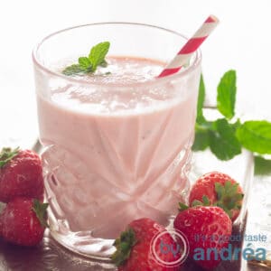 square photo with a glass filled with a strawberry orange yogurt smoothie, a red straw and surrounded with fresh strawberries and mint