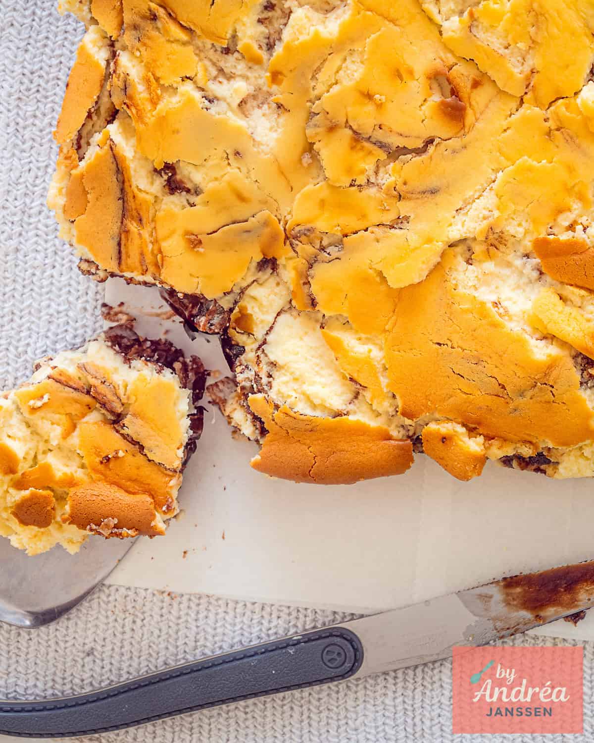 Een foto van bovenaf waar een cheesecake brownie op een bakpapier ligt en er een stuk is afgesneden.