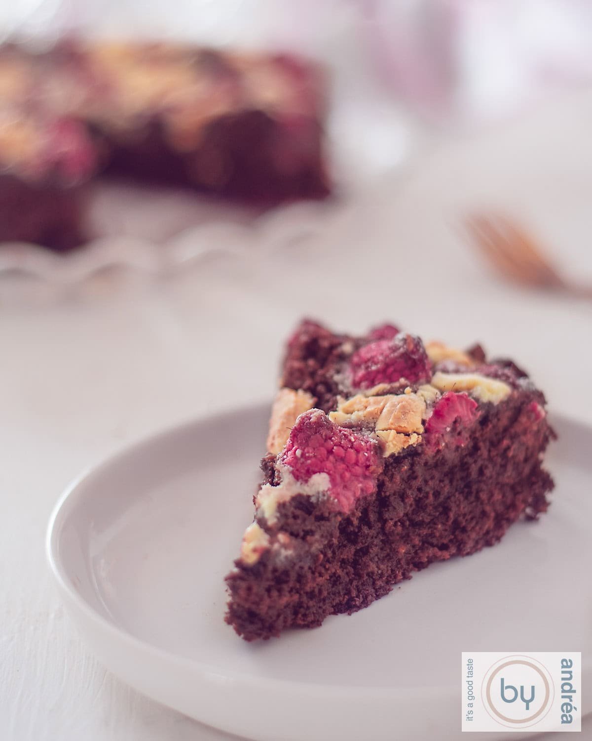 A slice of chocolate brownie with white chocolate chips and raspberries on a white plate. The rest of the cake is in the background.