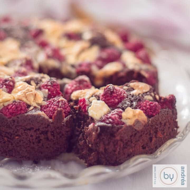 A square photo of a brownie with a slice cut out of it. A topping of white chocolate and raspberries.