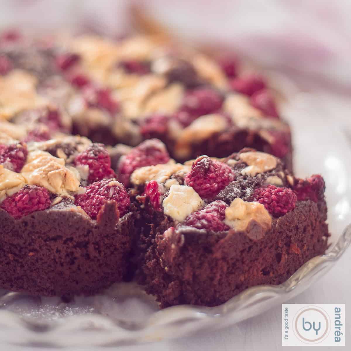 A square photo of a brownie with a slice cut out of it. A topping of white chocolate and raspberries.
