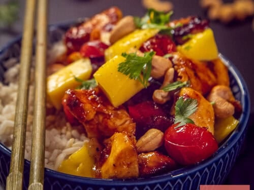 A square photo of a blue bowl with rice, mango topping, chicken in honey ketjap sauce, cashews and tomatoes.