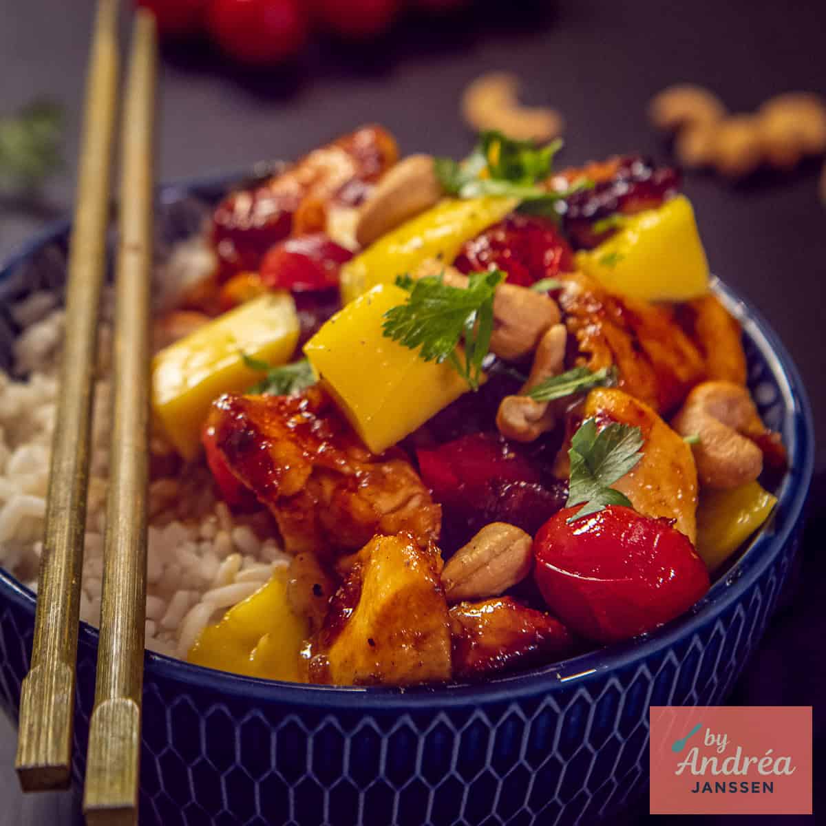 A square photo of a blue bowl with rice, mango topping, chicken in honey ketjap sauce, cashews and tomatoes.
