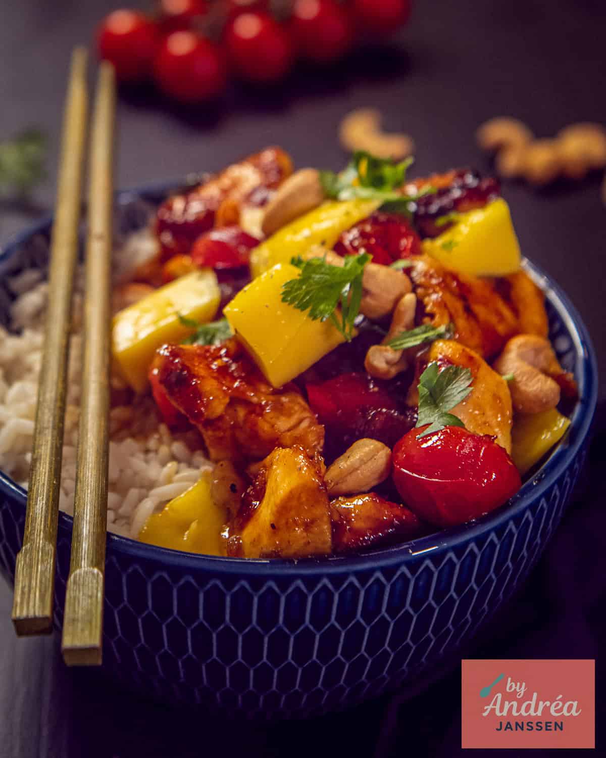 A blue bowl with honey ketjap chicken with sauce, tomato, mango and cashew nuts.