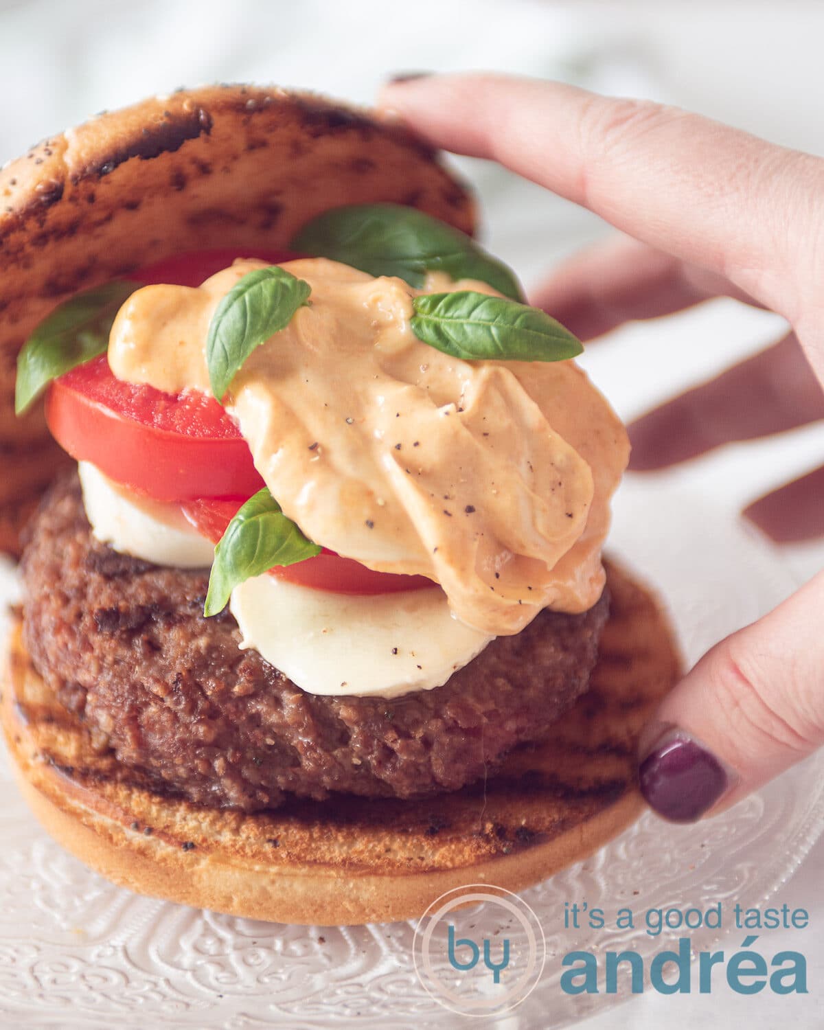 a hand taking a hamburger bun containing a juicy hamburger, mozzarella slices, tomato and pesto mayonaise