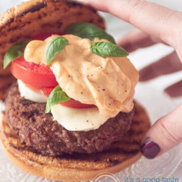 a hand taking a hamburger bun containing a juicy hamburger, mozzarella slices, tomato and pesto mayonaise