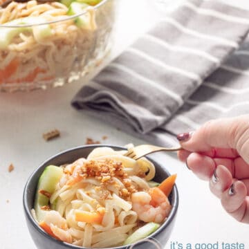 a hand taking a bit of the noodle salad in a great bowl. A big bowl filled with salad and a grey towel are in the back