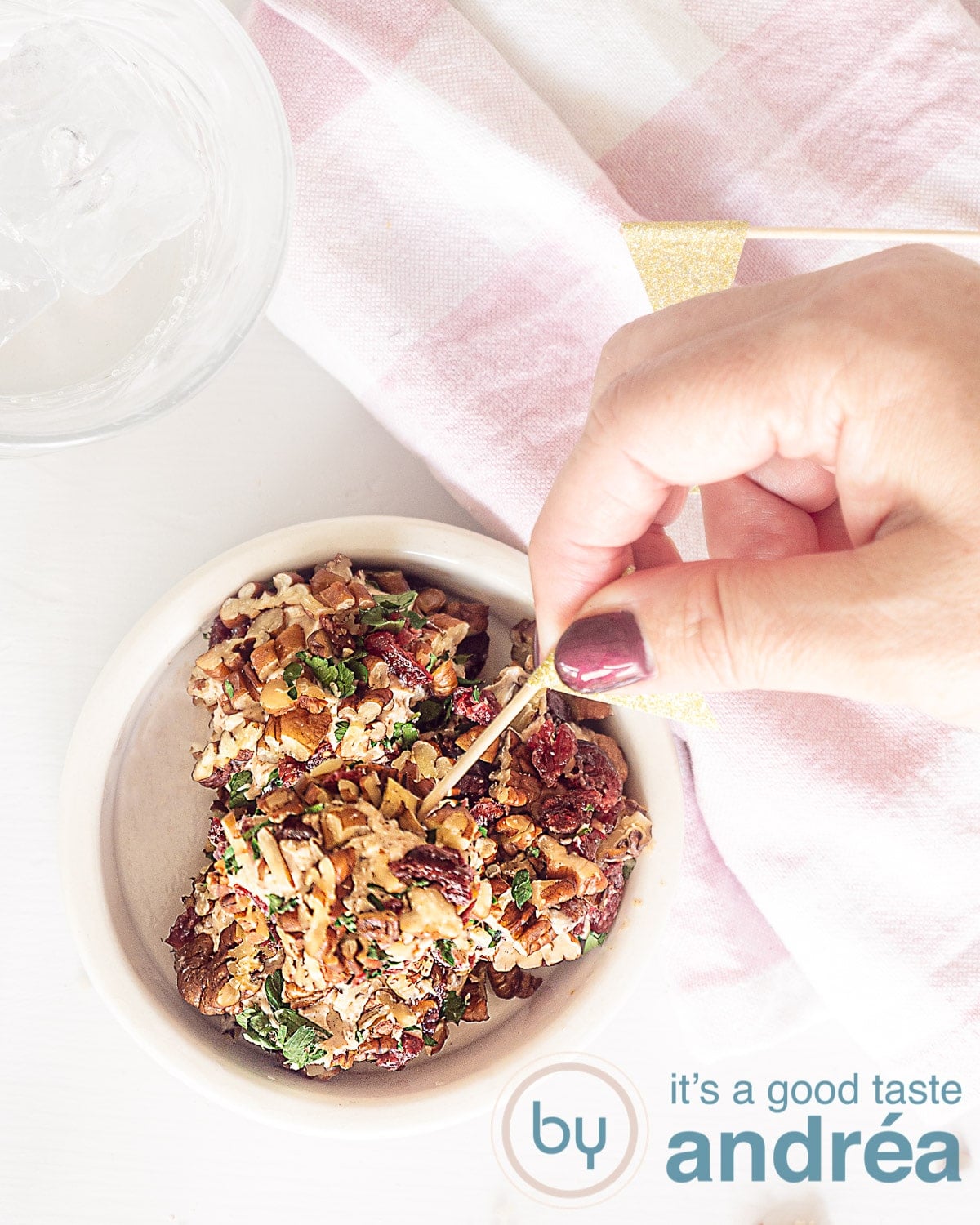A hand that uses a toothpick to remove a goat's cheese amuse-bouche ball from a white bowl. A pink and white checkered towel in the top corner