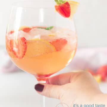 A hand holding a glass of strawberry gin cocktail with a strawberry and lemon garnish