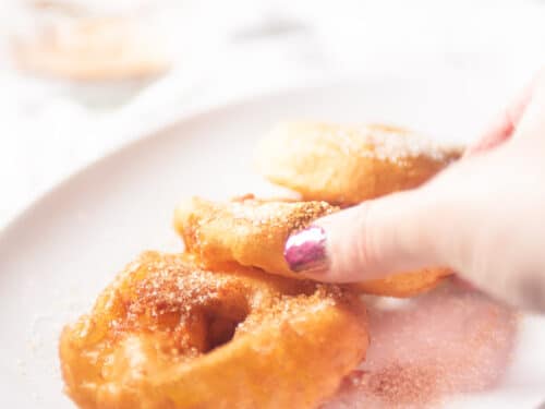 A hand taking an apple fritter from a white bowl with three fritters