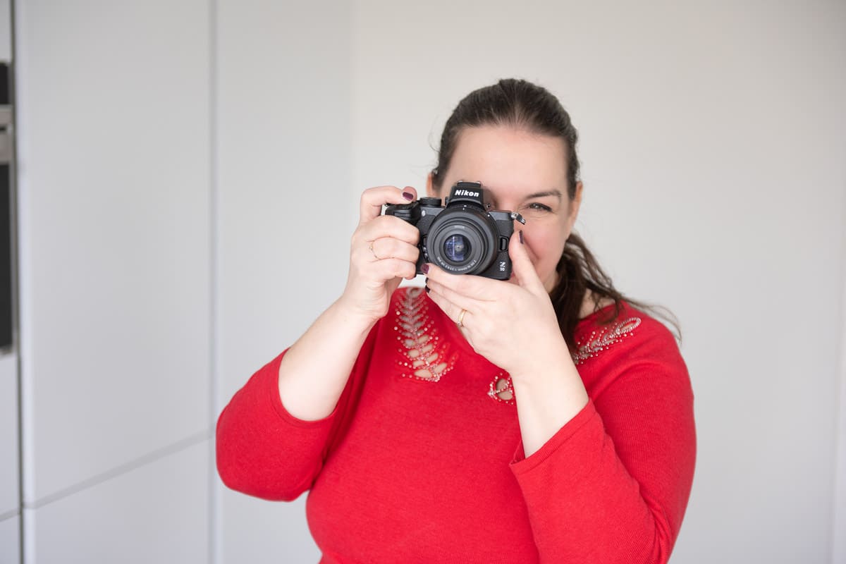 Andréa in the middle of the photo wearing a red sweater. Holding a camera in shooting position. A white background