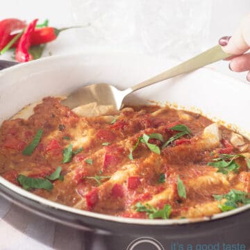 A hand holding a spoon that wants to grab a mackerel from the baking dish. The oven dish is black on the outside. Some peppers in the background