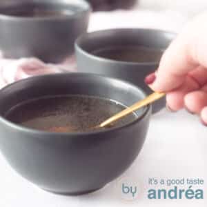 A square photo with gray bowls of chicken stock. A hand with a golden spoon takes a bite.
