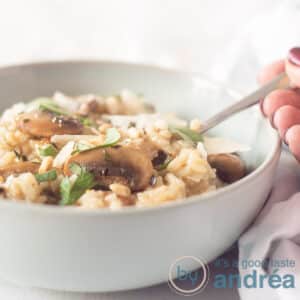 A blue dish with risotto, chestnut mushrooms and herbs. A hand takes a spoon. A white background