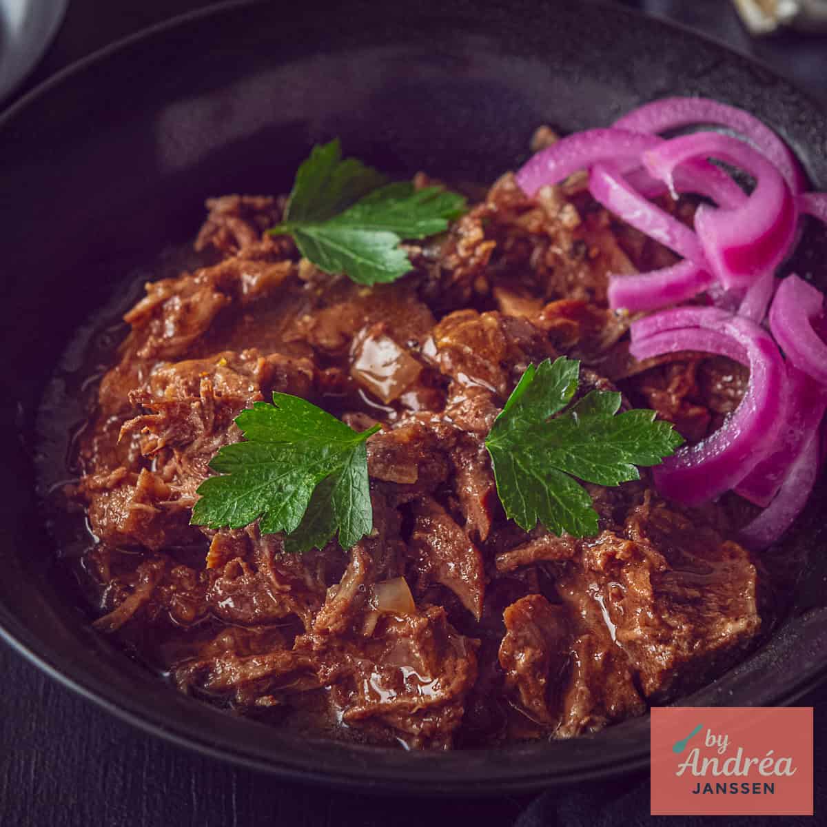 A square picture with pulled beef, parsley and onions in a black bowl.