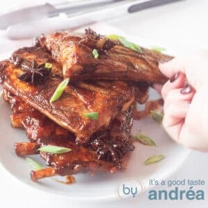 A square photo with a stack of sticky spareribs on top of each other. Sprinkled with spring onion. A hand takes one from the bowl