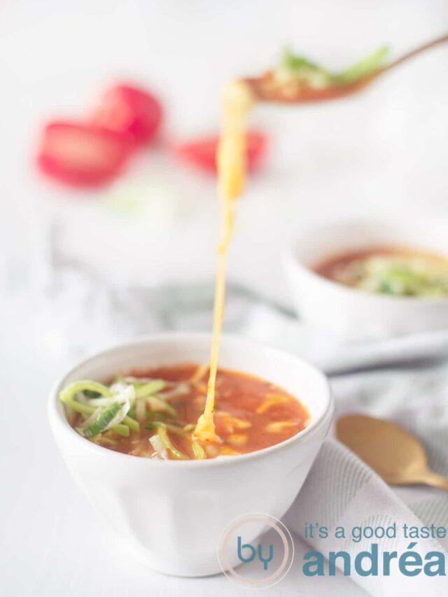 Two bowls of tomato soup (one front and center) with leeks and cheese. A spoon takes a bite and melted cheese hangs between the soup and the spoon. A green and white tea towel lies between the bowls with two golden spoons on top. diced tomatoes in the background