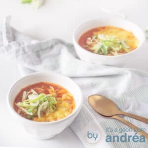 A square photo of a white background with two white bowls filled with tomato soup. With a topping of leek and cheese. Two golden spoons at the bottom left